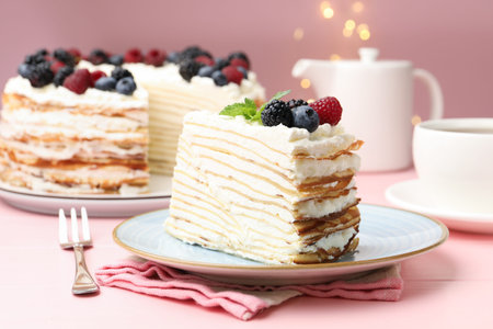Piece of tasty crepe cake with berries served on pink wooden table against blurred lights, closeup