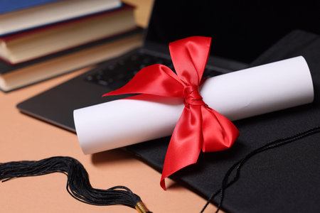 Graduation cap, diploma, laptop and books on beige table, closeup