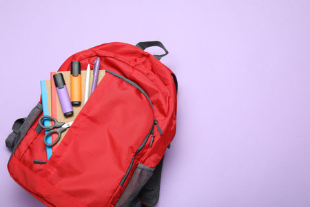 Red backpack with different stationery on violet background, top view. Space for text