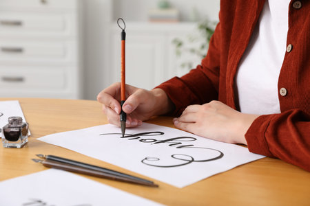 Calligraphy. Woman with brush writing word Enjoy on sheet of paper at wooden table, closeup