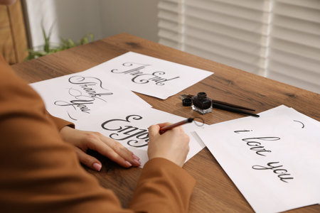 Calligraphy. Woman with brush writing word Enjoy on sheet of paper at wooden table, closeup