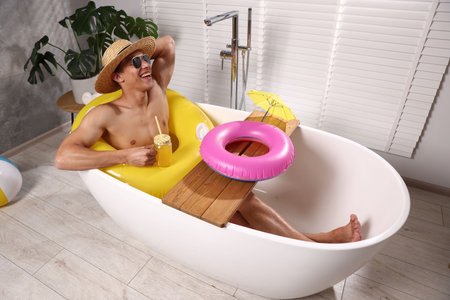 Happy man in sunglasses and straw hat drinking juice from mason jar with inflatable ring in bathtub...