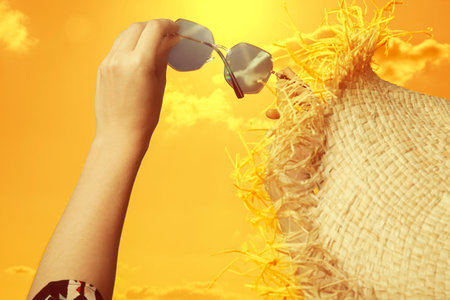 Woman holding sunglasses against orange sky with bright sun, closeup