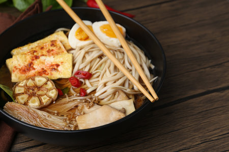 Tasty ramen with enoki and king oyster (eryngii) mushrooms in bowl on wooden table, closeup