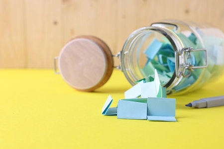 Colorful paper notes in glass jar and marker on yellow table, closeup. Space for text