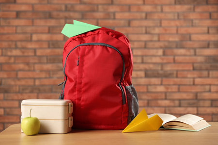 Backpack, paper planes, book, lunchbox and apple on wooden table against red brick wall