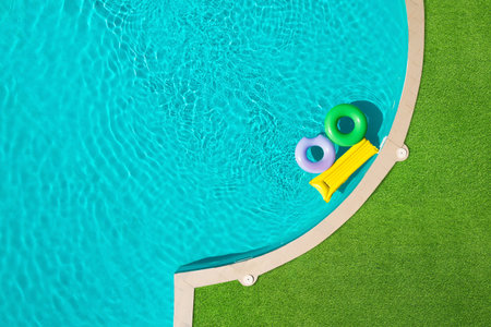 Inflatable rings and mattress floating in swimming pool surrounded by green grass, top view