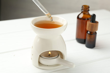Dripping essential oil from pipette into aroma lamp with burning candle on white wooden table...
