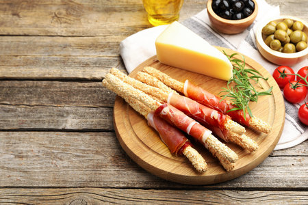 Delicious grissini sticks with sesame seeds, prosciutto and other products on wooden table, closeup....