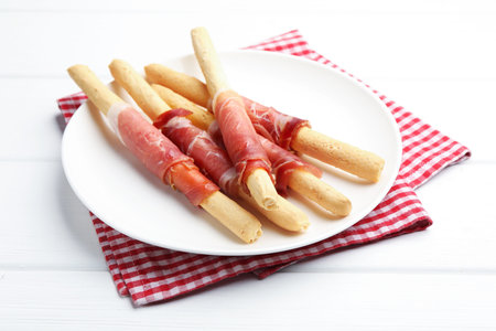 Delicious grissini sticks with prosciutto on white wooden table, closeup