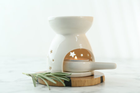 Aroma lamp with burning candle and rosemary on white marble table, closeup