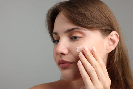 Beautiful woman applying cream onto her face on gray background, closeup. Space for textの写真素材