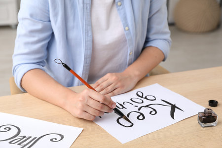Calligraphy. Woman with brush writing phrase Keep Going on sheet of paper at wooden table indoors,...