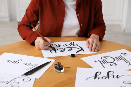 Calligraphy. Woman with brush writing phrase Keep Going on sheet of paper at wooden table indoors,...