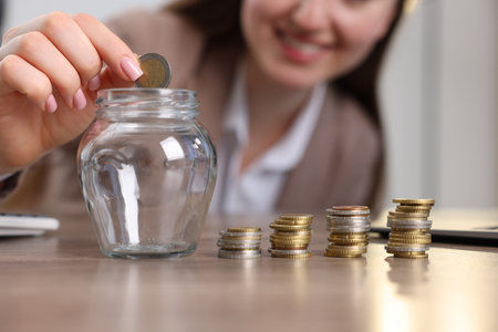 Woman putting coin into glass jar at table indoors, closeupの写真素材