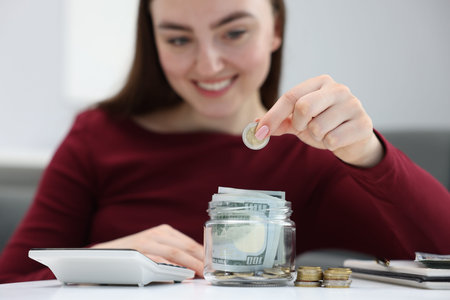 Woman putting coin into glass jar at table indoors, selective focus