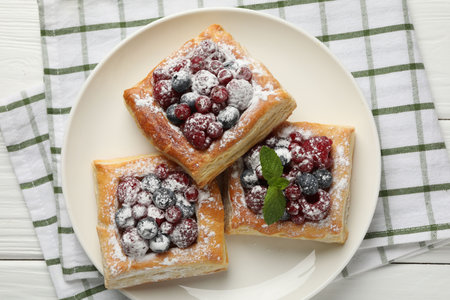Tasty puff pastries with berries, powdered sugar and mint on white wooden table, top view
