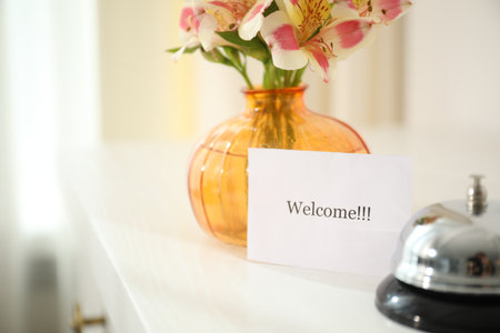 Card with Welcome word, desk bell and vase with flowers on white table indoors, closeupの写真素材