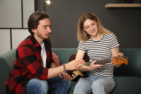 Tutor teaching woman to play guitar on sofa at home