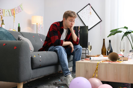 Man suffering from hangover on sofa in messy room after partyの写真素材