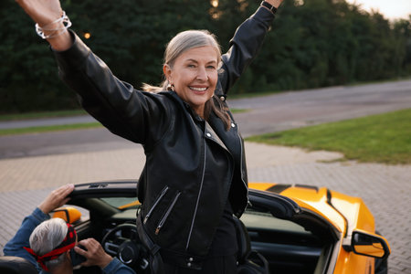 Senior couple in modern cabriolet outdoors. Woman with wide open arms and man talking on phone