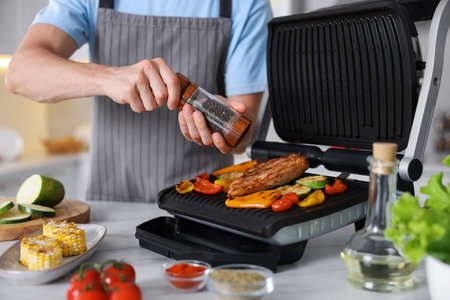 Man adding pepper to vegetables and meat cooking on electric grill in kitchen, closeup