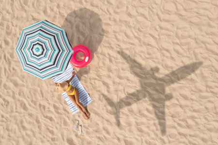 Woman resting under striped umbrella on beach, top view. Flying airplane casting shadow on sandの写真素材
