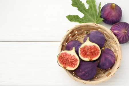Fresh ripe figs in wicker basket and green leaf on white table, flat lay. Space for textの写真素材