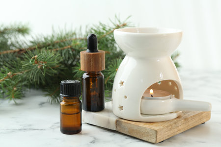 Aroma lamp with burning candle, bottles of essential oils and fir tree branches on white marble...