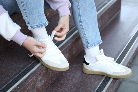 Woman tying shoelace of white sneaker on stairs outdoors, closeup
