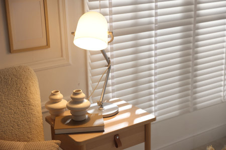 Stylish lamp, vases and book on wooden bedside table near armchair indoors