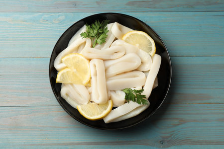 Raw squid rings, parsley and lemon in bowl on light blue wooden table, top view