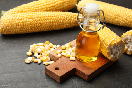 Corn oil in glass bottle, kernels and cobs on gray textured table, closeup