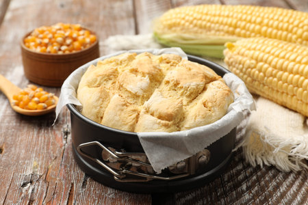 Freshly baked cornbread in cake pan, cobs and napkin on color wooden table, closeup