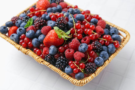 Different ripe berries in wicker basket on white tiled table, closeup