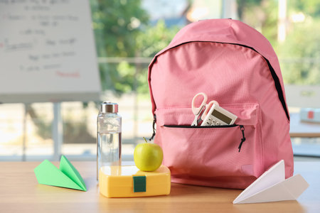 Backpack with different stationery, lunchbox, apple, bottle of water and paper planes on desk in...