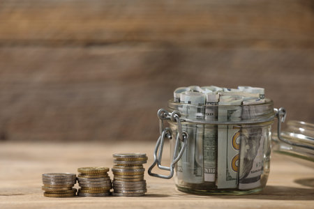 Glass jar with dollars and coins on wooden table, closeup