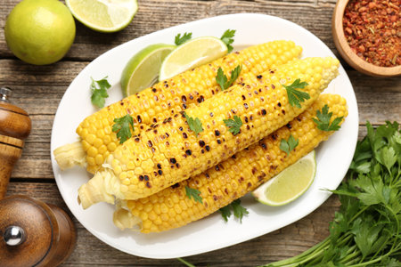 Tasty grilled corncobs with parsley and lime slices on wooden table, flat lay