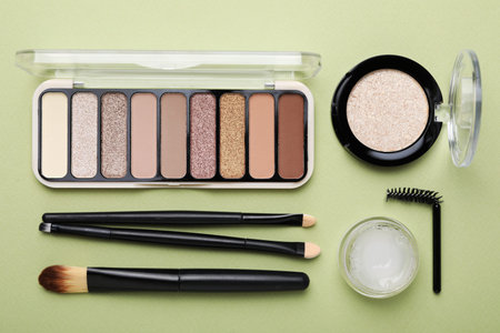 Colorful eyeshadow palette, other cosmetic products, makeup applicators and brushes on green...