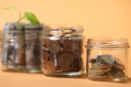 Glass jars with coins and sprout on pale orange background, closeup