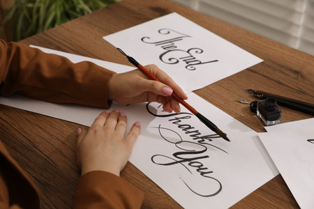 Calligraphy. Woman with brush writing words Thank You on sheet of paper at wooden table, closeup
