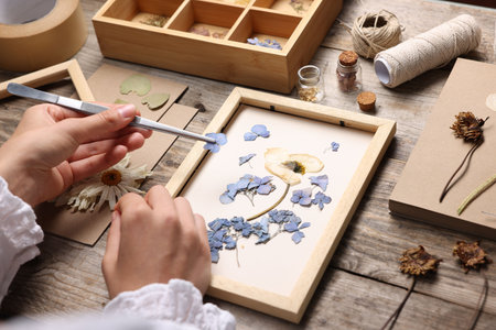 Woman putting dry flowers into picture frame at wooden table, closeup