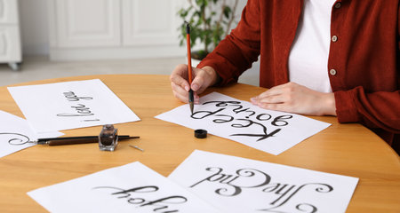 Calligraphy. Woman with brush writing phrase Keep Going on sheet of paper at wooden table indoors,...