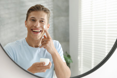 Handsome young man applying cream onto his face near mirror at homeの写真素材