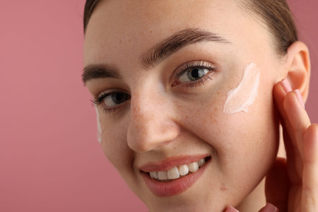 Beautiful woman with cream on her face against pink background, closeup