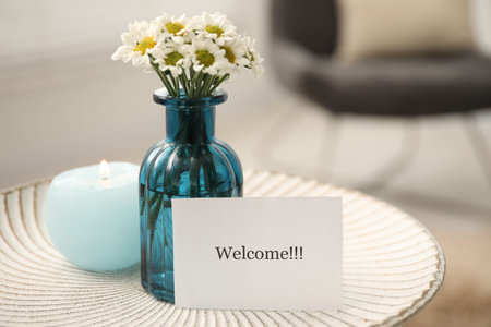 Card with Welcome word, burning candle and vase with flowers on white table indoors, closeup. Space...