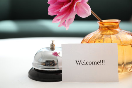 Card with Welcome word, desk bell and vase with flower on white table indoors, closeup