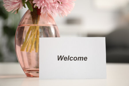 Card with Welcome word and vase with flowers on white table indoors, closeupの写真素材