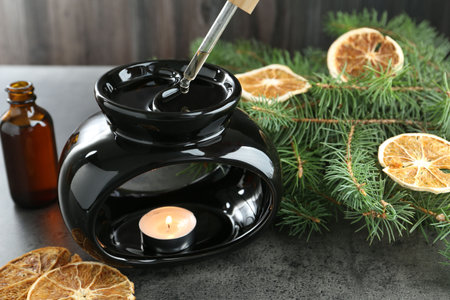 Dripping essential oil from pipette into aroma lamp with burning candle at gray textured table...