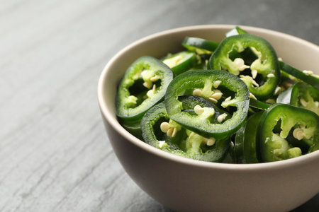 Pieces of green jalapeno peppers in bowl on gray table, closeup. Space for text